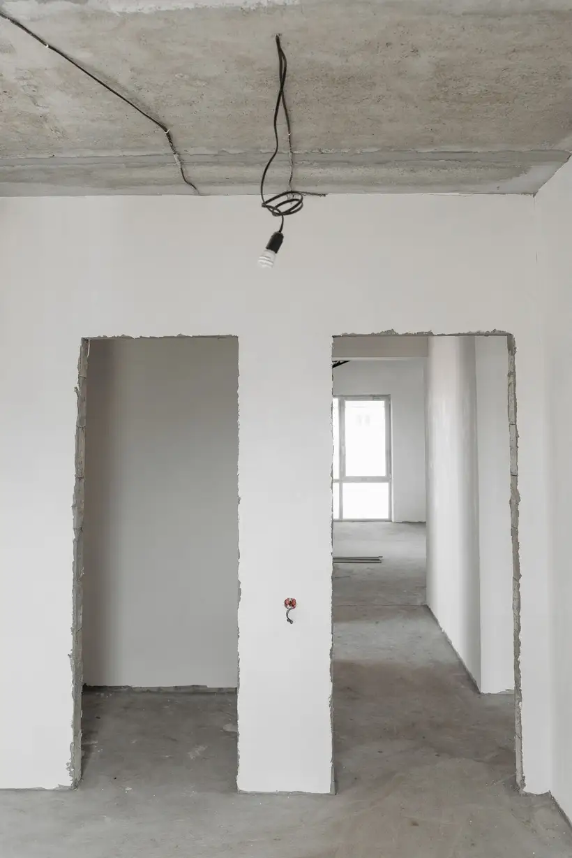 Surowe wnętrze po robotach budowlanych: betonowy sufit z odsłoniętym okablowaniem, otynkowane ściany działowe bez wykończenia i posadzka z betonu.