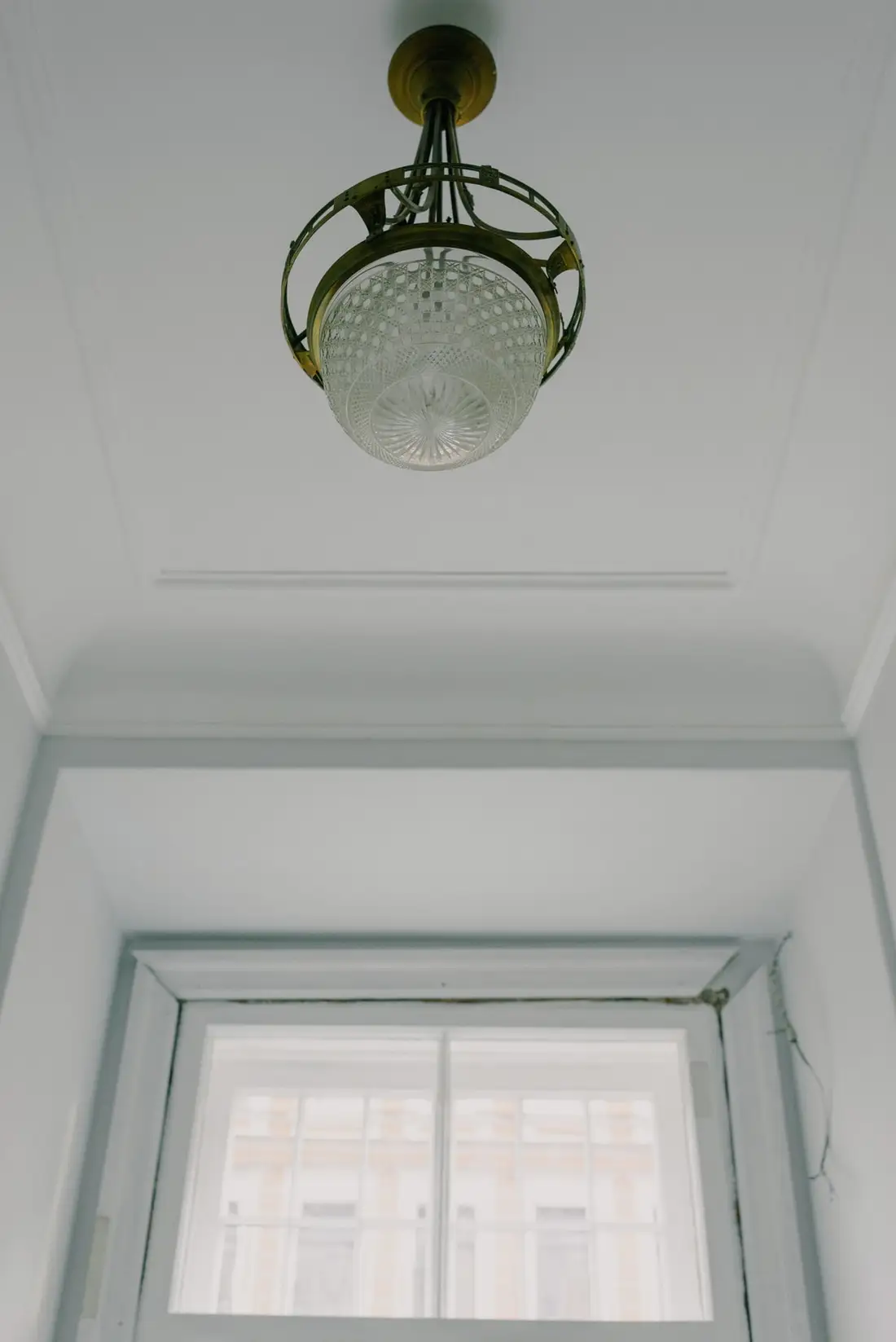 Zabytkowa mosiężna lampa sufitowa z ryflowanym kloszem, zawieszona pod wysokim, sztukatorskim sufitem nad oknem kamienicy.